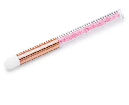 A close-up image of a lash and brow cleaning brush with a transparent handle containing pink crystal particles, featuring a white bristle head and a rose gold-colored ferrule.